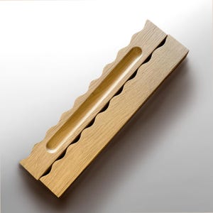 Puede incluir: Un porta-inciensos de madera clara que presenta un canal largo, estrecho y hundido para varitas de incienso. El objeto tiene un borde ondulado y festoneado distintivo en un lado, mostrando la veta natural y la textura de la madera.