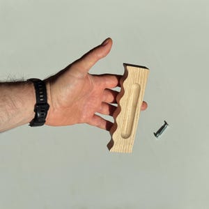 Könnte beinhalten: Ein heller Holz-Möbelgriff mit einer gewellten, geschwungenen Kante an einer Seite und einer vertieften, länglich ovalen Griffmulde in der Mitte. Der Griff ist rechteckig geformt, hat eine glatte Oberfläche und enthält eine Metallschraube.