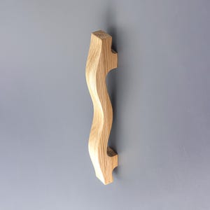 May include: A vertical cabinet handle crafted from light-colored natural wood with a distinct wavy, serpentine silhouette. The handle features a visible wood grain pattern and a smooth finish, mounted against a solid, flat, matte gray surface.