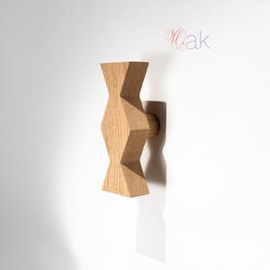 May include: A light brown wooden wall hook with a geometric, hourglass-like shape featuring faceted edges and a cylindrical base. The word Oak is printed in a stylized, thin font in the upper right corner of the image.