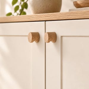 May include: Two circular light-coloured wooden cabinet knobs are mounted on a pair of off-white cabinet doors. The knobs feature a natural wood grain texture and a cylindrical shape with a small cutout detail on the underside of each handle.