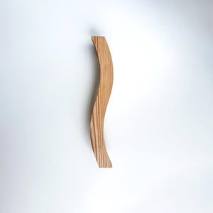 Może przedstawiać: Pionowy drewniany uchwyt o wyraźnym zakrzywieniu w kształcie litery S. Przedmiot wykonany jest z jasnego drewna z widocznym liniowym wzorem słojów biegnącym wzdłuż całej długości. Uchwyt posiada gładką powierzchnię oraz jednolitą grubość.