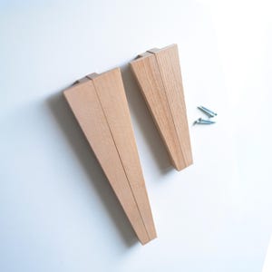May include: Two unfinished, light-colored wooden furniture legs with a triangular design. The legs are positioned on a white surface, with two small silver screws nearby. The legs are made of wood and are ready for finishing.