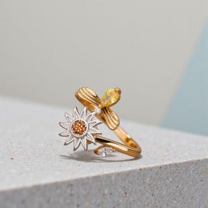 Puede incluir: Anillo dorado con diseño de girasol y abeja. El girasol tiene una capa exterior plateada y un centro de pequeñas piedras anaranjadas. La abeja presenta una gema amarilla. El diseño del anillo es delicado e inspirado en la naturaleza.