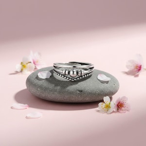 May include: A silver ring with a layered design, featuring a central beaded element and a delicate, adjustable band. The ring sits on a gray stone, surrounded by pink flower petals against a soft pink background.