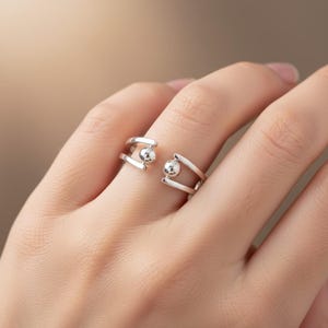 Könnte beinhalten: Ein silberfarbener offener Ring mit minimalistischem Design. Der Ring hat zwei parallele Bänder, die durch eine kleine, kugelförmige Perle auf jeder Seite verbunden sind. Der Ring wird an einem Finger getragen und zeigt seine moderne und schlichte Ästhetik.