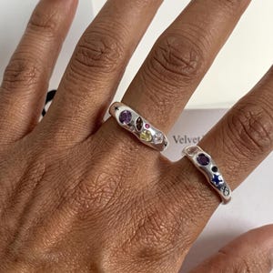 Pode incluir: Dois anéis de prata com bandas irregulares e orgânicas incrustadas com várias pedras preciosas multicoloridas e pequenos motivos de estrelas gravados. As pedras incluem gemas facetadas roxas, amarelas e claras, além de uma estrela azul incrustada.
