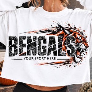 Może przedstawiać: Biała bluza z długim rękawem i krótkim krojem, ozdobiona grafiką głowy tygrysa oraz napisem BENGALS YOUR SPORT HERE czcionką w stylu postarzanym, z pomarańczowymi akcentami i efektem rozpryskanej farby na przedniej części ubrania.