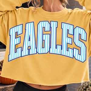 Peut inclure: Un sweat-shirt court à manches longues jaune moutarde avec le mot EAGLES imprimé en grandes lettres arquées. Les lettres sont bleu clair avec des rayures verticales blanches et un contour bleu foncé sur le tissu jaune uni.