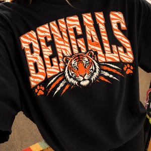 May include: A black t-shirt featuring the word BENGALS in large, orange and white tiger-striped block letters. Below the text is a detailed illustration of a tiger face with orange, black, and white fur, flanked by two orange paw prints.