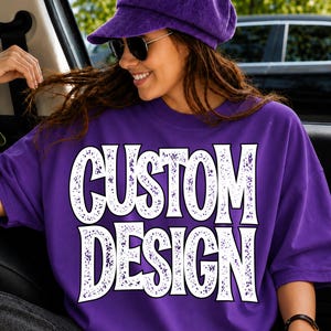 May include: A purple corduroy newsboy cap and a purple short-sleeve t-shirt featuring the text CUSTOM DESIGN in large, white, distressed-effect block letters across the chest. The shirt has a crew neckline and a loose, relaxed fit.