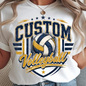Peut inclure: Un t-shirt blanc avec un motif graphique comprenant le mot CUSTOM en lettres capitales bleu marine usées au-dessus d'une illustration de ballon de volley. Le mot Volleyball est écrit en jaune en dessous, avec des détails bleu marine et jaune.