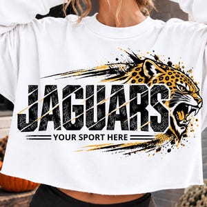 Op de afbeelding: Een wit, kort sweatshirt met lange mouwen en een afbeelding van een grommende jaguarkop in geel en zwart. De tekst JAGUARS is gedrukt in grote zwarte letters met een verweerd effect, met daaronder de tekst YOUR SPORT HERE in het zwart.