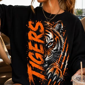 Puede incluir: Una sudadera negra de manga larga con un gran estampado gráfico de la cara y la pata de un tigre con garras afiladas. La palabra TIGERS está impresa verticalmente en letras naranjas desgastadas junto al tigre, que tiene pelaje naranja y blanco.