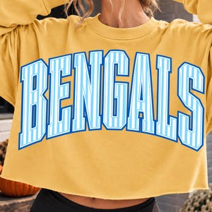 May include: A mustard yellow cropped sweatshirt featuring the word BENGALS printed in a large, arched font. The letters are light blue with white vertical stripes and a dark blue outline, set against the solid yellow fabric of the garment.