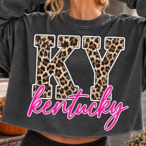 May include: A charcoal grey cropped sweatshirt featuring the letters KY in a brown and black leopard print pattern. Below the letters, the word Kentucky is printed in a bright pink script font with a white outline on the front of the garment.