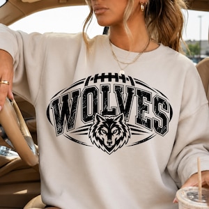 May include: A beige crewneck sweatshirt featuring a black graphic print of a football shape containing the text WOLVES in a bold, distressed font, centered above a stylized wolf head illustration with sharp, angular lines and details.