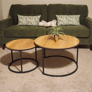 May include: Two round wooden coffee tables with black metal frames. The tables are different sizes and sit on a light-colored carpet. A green sofa with patterned pillows is in the background. A small plant in a gold pot sits on the larger table.
