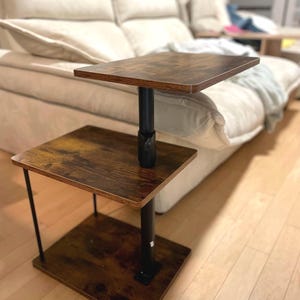 May include: A two-tiered side table with a dark brown, wood-grain finish. The table has a square top and a lower shelf, connected by a black, adjustable-height pole. The table is supported by a square base and black metal legs.