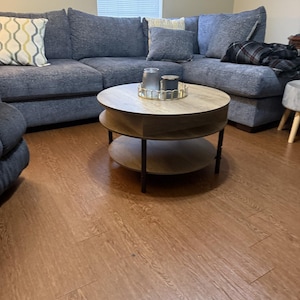 May include: A blue sectional sofa with decorative pillows and a round, two-tiered coffee table. The coffee table has a light wood top and a dark metal base. Two glass candle holders sit on a gold tray.