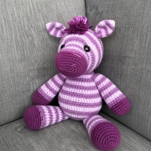 May include: A handmade crochet zebra stuffed animal in shades of purple and white. The zebra has a dark purple muzzle, hooves, and a tuft of hair. The body features alternating stripes of light purple and white. The toy is sitting on a gray surface.