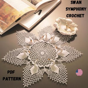 Puede incluir: Un tapete de ganchillo blanco con un diseño circular central y un patrón de estrella. Seis figuras de cisnes blancos están dispuestas alrededor del centro. El texto "SWAN SYMPHONY CROCHET" y "PDF PATTERN" son visibles. Un libro abierto está en el fondo.