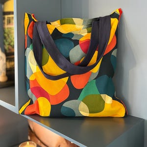 Op de afbeelding: Een kleurrijke totebag met een levendig abstract patroon van overlappende vormen in geel, oranje, rood, groen en teal op een donkere achtergrond. De tas heeft marineblauwe hengsels en ligt op een grijze plank.