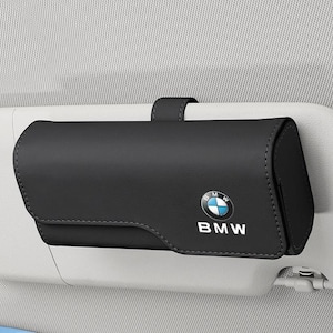 May include: A black rectangular car visor sunglasses holder featuring the BMW logo and text in white. The case is constructed with a smooth finish and includes a strap for secure attachment to a vehicle sun visor, providing storage for eyewear.