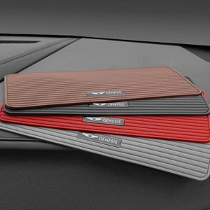 Könnte beinhalten: Ein Stapel von vier rechteckigen Auto-Armaturenbrettmatten in Braun, Schwarz, Rot und Grau. Jede Matte verfügt über eine horizontal gerippte Textur und ein zentrales Metallemblem mit dem Genesis-Logo und dem Schriftzug GENESIS in Weiß.