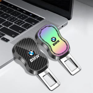 May include: Two metal seat belt buckle inserts featuring the BMW logo. One insert has a black carbon fiber pattern, while the other has a gradient iridescent finish with a decorative border. Both items are shaped with a rounded top and a rectangular slot.