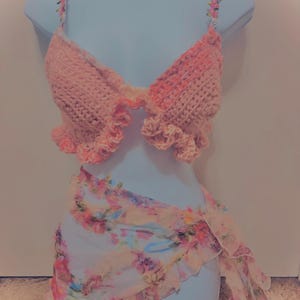 May include: A light pink crochet top with floral straps and ruffled edges. A matching floral print wrap skirt with ruffled trim is also included. The outfit is displayed on a mannequin.