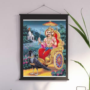 May include: A framed print depicting a deity in a golden chariot, holding a staff and umbrella. A black crow stands in front of the chariot. The background shows a temple, mountains, and a lake. The print is framed in black and hangs on a white wall.