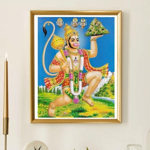 May include: A framed print of the Hindu deity Hanuman in a gold frame. The artwork shows Hanuman with a mace and holding a mountain. The background is a blue sky with a green and yellow landscape. The print is displayed on a white mantelpiece.
