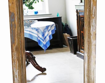 May include: Rectangular mirror with a distressed, weathered wooden frame. The frame has a mix of brown, tan, and white paint. The mirror reflects a room with a table, window, and other furniture.