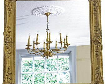 May include: A large, rectangular gold mirror with an ornate, carved frame. The mirror reflects a chandelier with multiple lights and a window with greenery. The frame has detailed scrollwork and floral designs.