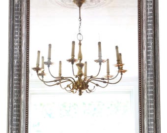 May include: A rectangular mirror with an arched top and a distressed silver frame. The mirror reflects a brass chandelier with multiple candle-shaped lights. The frame has a detailed, textured design. The overall style is vintage.
