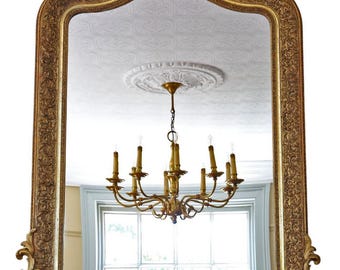 May include: Ornate gold-framed mirror with intricate carvings. The mirror reflects a chandelier and a window. The frame has a detailed, textured design, adding a touch of elegance. The mirror is a decorative home decor item.