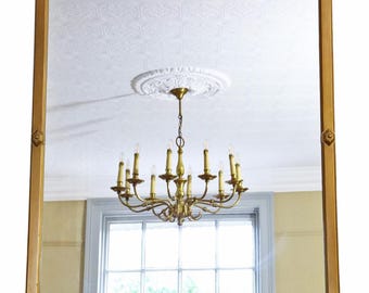 May include: A large, rectangular gold-framed mirror with decorative corner and top accents. The mirror reflects a chandelier with multiple candle-shaped lights and a window. The frame has a simple, elegant design.