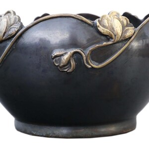 May include: A dark bronze-colored decorative bowl with a scalloped rim. The bowl features gold-colored floral accents and a rounded shape. The floral design is repeated around the rim of the bowl, adding a touch of elegance.