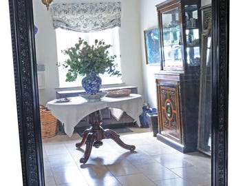 May include: A large, rectangular black framed mirror with ornate detailing. The mirror reflects a room with a round table, a vase of flowers, and a cabinet. The frame has a detailed floral pattern.