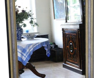 May include: Rectangular mirror with a gold and silver frame. The mirror reflects a room with a table, vase, and cabinet. The frame has a black outer edge and a rounded top.