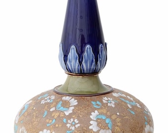 Op de afbeelding: Een decoratieve keramische vaas met een bolvormige basis en een lange, slanke hals. De basis heeft een bruinachtige achtergrond met gouden en turquoise bloempatronen. De hals is donkerblauw en de bovenkant is groen.