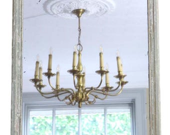 May include: A rectangular mirror with a distressed, light gray frame. The mirror reflects a brass chandelier with multiple candle-shaped lights, a white ceiling medallion, and a window with greenery.