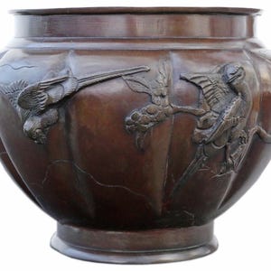 May include: A large, antique bronze planter with a wide, fluted body. The planter features detailed relief carvings of birds and foliage. The bronze has a rich, dark patina, and the planter sits on a low, circular base.