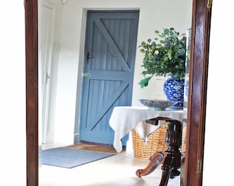 May include: A large, rectangular, antique mirror with a dark wood frame. The top of the mirror has an arched design with gold accents. The mirror reflects a room with a blue door and a table with a vase.