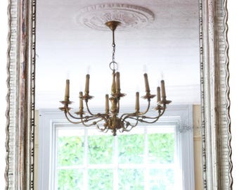 May include: A rectangular mirror with a distressed, ornate silver frame. The mirror reflects a chandelier with multiple candle-style lights and a window with greenery. The frame has a wavy edge.