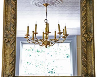 May include: A large, rectangular mirror with an ornate, gold-colored frame. The mirror reflects a room with a chandelier and a window. The frame features detailed carvings and a textured surface, adding a vintage aesthetic.