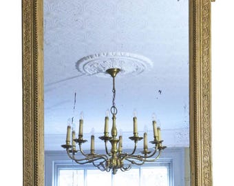 May include: A rectangular gold-framed mirror with ornate detailing. The top of the frame features a decorative crest with floral and leaf motifs. The mirror reflects a room with a chandelier and a window.