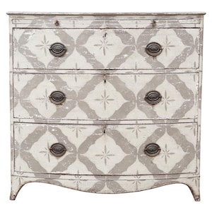 May include: A three-drawer chest of drawers with a distressed, painted finish. The chest is painted white with a gray geometric pattern. It has antique-style metal drawer pulls and keyholes.