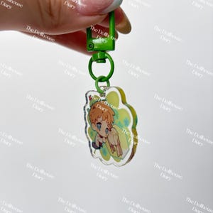 May include: A clear acrylic keychain with a green clasp and ring. The keychain features a cartoon illustration of a character with orange hair and blue eyes. The background is a light green floral design. The text "The Dollhouse Diary" is printed on the keychain.